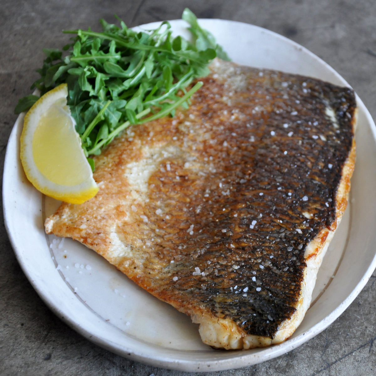 Seared fish fillet on a plate with lemon and greens on a textured surface